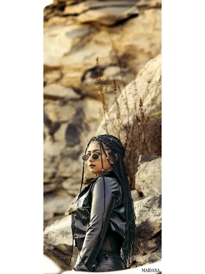 A vertically composed shot of the model with braided hair and sunglasses, looking back against a rocky backdrop. The unique framing makes this a visually interesting and edgy portrait.