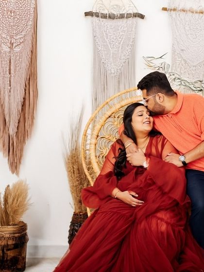 A loving embrace in the studio. The partner kisses the mother-to-be's forehead as she sits in a wicker chair, surrounded by a cozy, rustic setup.