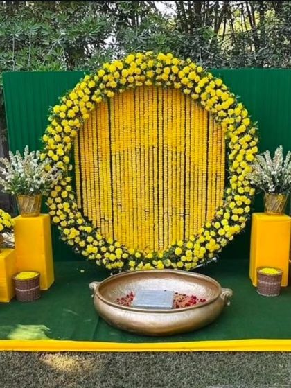 A classic Haldi backdrop for outdoor functions. This setup features a large circular frame filled with strings of yellow marigolds, a green panel background, and a traditional brass urli in the center for the ceremony.
