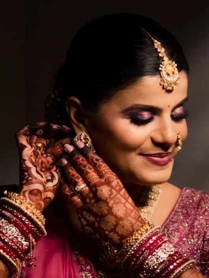 A classic bridal portrait. The bride is adjusting her earring, giving a beautiful view of her intricate henna, smokey purple eyeshadow, and the detailed embroidery on her pink lehenga.
