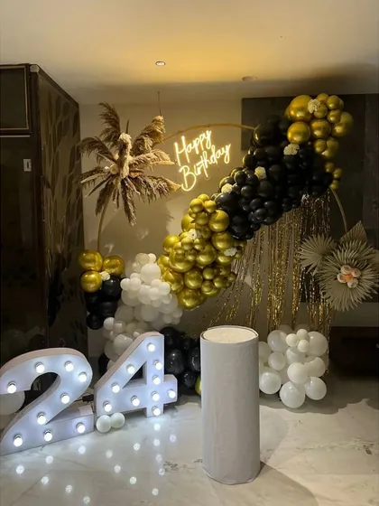 A chic 24th birthday setup with a black, white, and gold color scheme. This decoration includes a balloon garland, a neon sign, marquee numbers, and dried pampas grass for a boho touch.