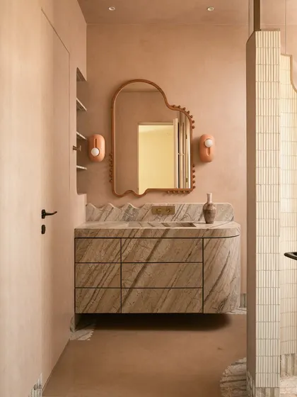 We call this bathroom a 'chic desert dream'. It features blush-toned micro-concrete walls and a monolithic vanity carved from veined Corteccia marble. The stone appears to trickle down onto the floor, creating a fluted skirting detail that is both organic and luxurious.