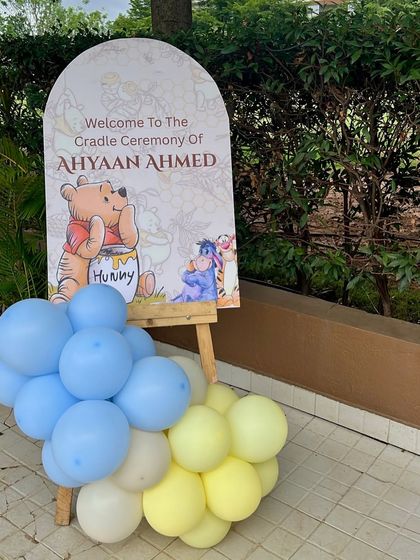 The welcome sign for Ahyaan's cradle ceremony, featuring a classic Winnie the Pooh illustration and a matching blue and yellow balloon cluster.