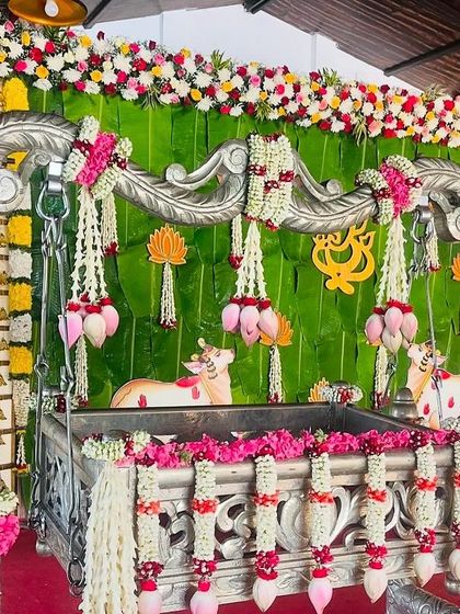 A beautifully decorated silver cradle for a naming ceremony. The setup includes a banana leaf backdrop, lotus hangings, and garlands made of jasmine and pink flowers.