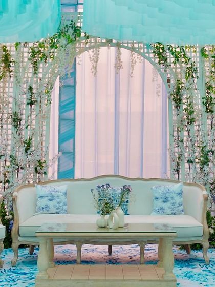 A chic seating area designed for a couture Mehendi. The lounge features elegant French-style furniture, custom-printed cushions, and a backdrop of white trellises with delicate floral arrangements.