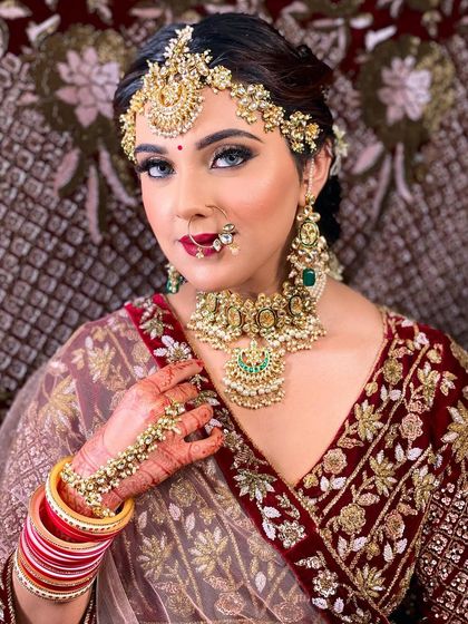 A stunning bridal portrait showcasing a classic look with a maroon lehenga and heavy kundan jewelry. The makeup features bold, defined eyes and a perfectly matched lip color, creating a regal and timeless appearance.