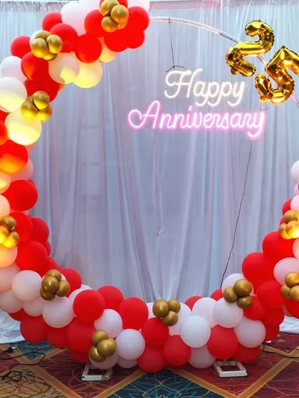 We design vibrant and cheerful anniversary decorations perfect for any space. This setup features a circular ring frame decorated with a full balloon arch in red, white, and gold, a 'Happy Anniversary' neon sign, and foil number balloons to mark the special milestone.