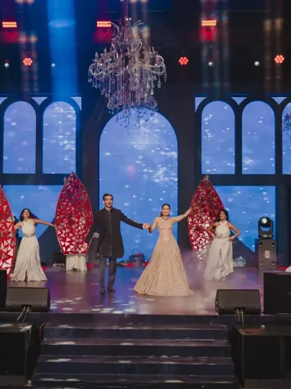 The couple takes center stage for their performance, framed by dramatic red set pieces and professional backup dancers, showcasing a perfectly choreographed Sangeet moment.