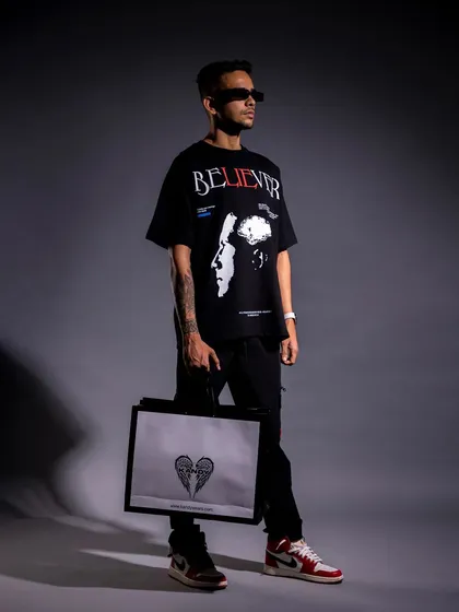 A full-length studio shot of a male model in an oversized graphic tee and sneakers, holding a branded bag. This is a complete look that's perfect for a lookbook, showing how different brand items can be styled together.