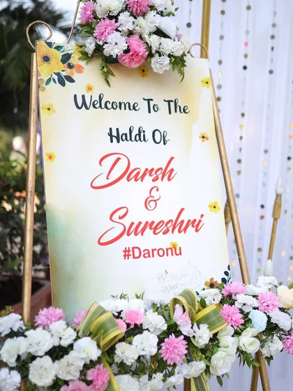 A personalized welcome sign for the Haldi ceremony of Darsh and Sureshi. The sign is beautifully decorated with floral arrangements, setting a warm and welcoming tone for the event.