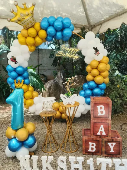 An outdoor first birthday celebration with a royal theme, featuring a blue, yellow, and white balloon arch topped with a crown.