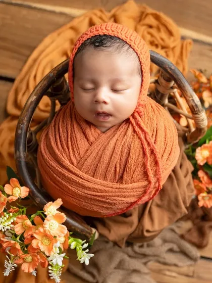 This newborn is wrapped in a warm orange swaddle and placed in a basket, surrounded by autumnal flowers for a cozy, seasonal feel.