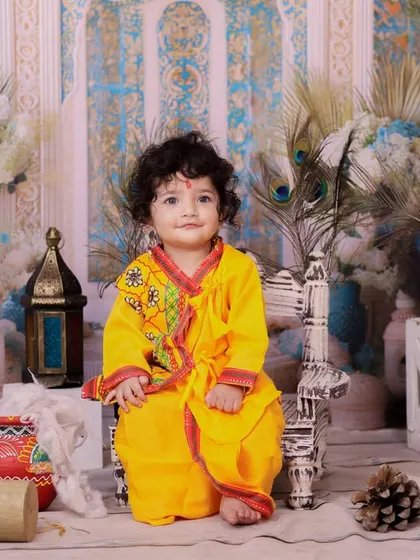 A baby dressed in a yellow kurta sits calmly on a small chair in our Janmashtami setup. The peacock feathers and painted pots add a traditional touch.