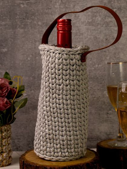 A gray crocheted wine bag is styled with glasses of champagne against a moody, textured backdrop. This shot creates a sophisticated and celebratory atmosphere, perfect for gifting.