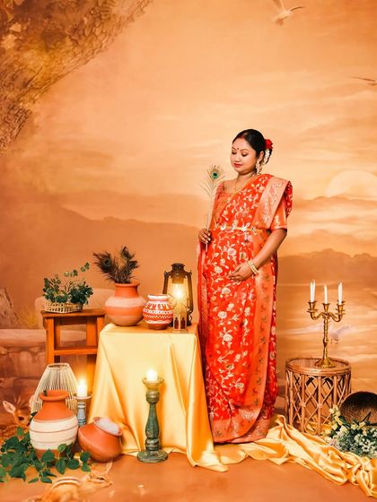 A beautiful solo portrait of a mom-to-be in a traditional red Banarasi saree. The backdrop, depicting a serene sunset over a river, adds a painterly and timeless quality to the image.