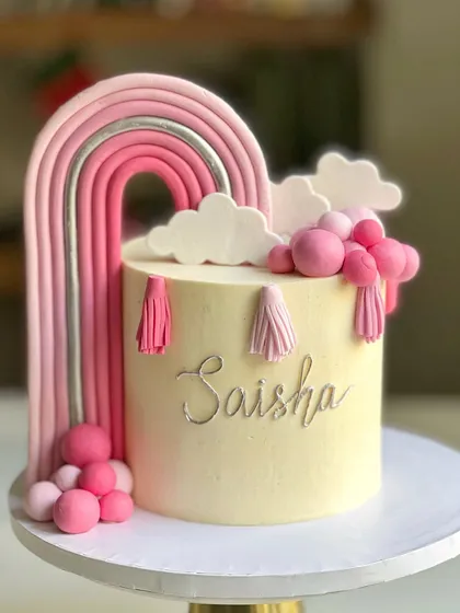 A boho-chic rainbow cake in shades of pink and silver. The fondant rainbow is accented with tassels and fluffy clouds, creating a trendy and stylish look for a birthday celebration.