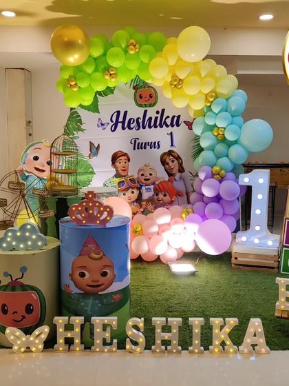 A Cocomelon-themed first birthday with a faux grass floor. The decor includes the iconic yellow bus, character cutouts, and a 'HESHIKA' marquee.