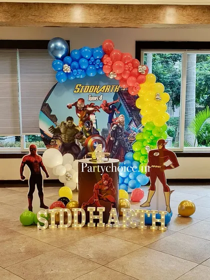 A powerful superhero-themed birthday party featuring the Avengers. The decor includes a dynamic backdrop, character cutouts of Spider-Man and The Flash, and a colorful balloon arch.