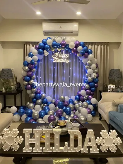 A beautiful balloon ring in blue, purple, and silver, set up in a living room. The fairy lights woven into the balloons and the light-up name sign create a magical, frozen-like effect.