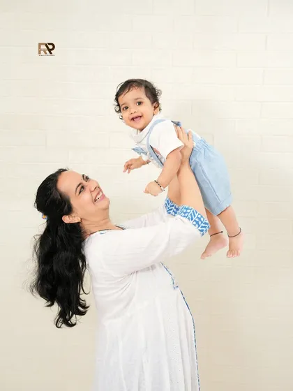 A pure moment of joy between a mother and her child. The simple white brick background keeps the focus on her happy expression as she lifts her baby in the air.