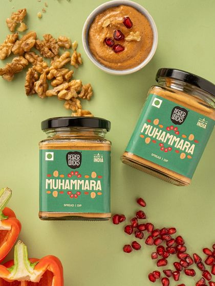 A clean and simple product shot of Muhammara dip. The ingredients, walnuts and pomegranates, are scattered around the jars, providing context and a fresh, natural feel. This is effective for both e-commerce and social media.