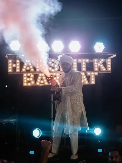 The groom makes a grand entrance for his baraat, using a smoke gun to amp up the energy. The custom 'Harshit ki Baraat' sign adds a personal and fun touch.