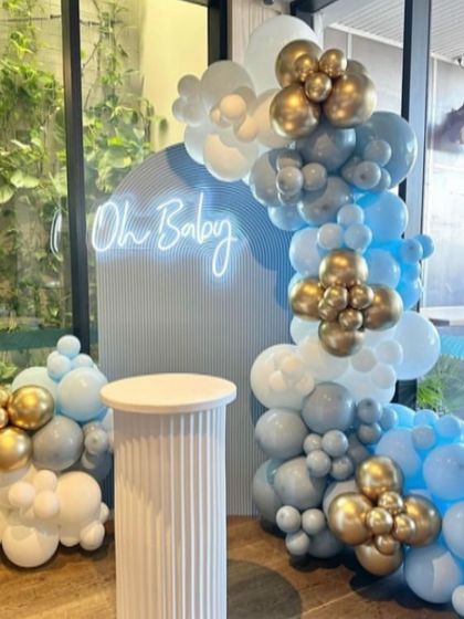 A modern baby shower setup with a blue arched backdrop and an 'Oh Baby' neon sign. The organic balloon garland uses a sophisticated palette of blue, silver, and white.