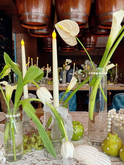 A fresh and elegant centerpiece combining white calla lilies and tulips with pearl-studded pears and green grapes. The mix of flowers and fruit on a lace runner creates a feeling of natural, effortless beauty.