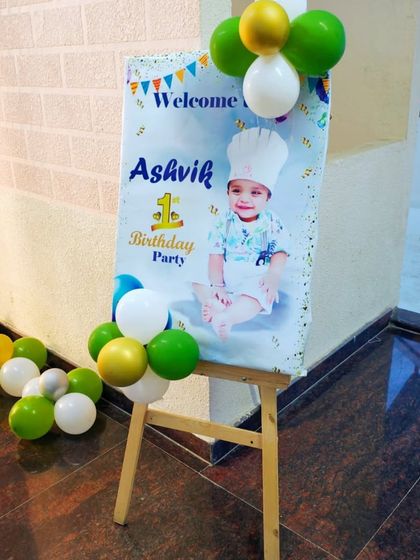 A custom welcome board for a first birthday party, complete with the baby's photo. The easel is decorated with a small balloon cluster that matches the party's color scheme.