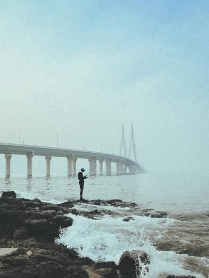 A lone fisherman stands on the rocks near the Bandra-Worli Sea Link, casting his line into the water. A powerful silhouette against the misty sea.