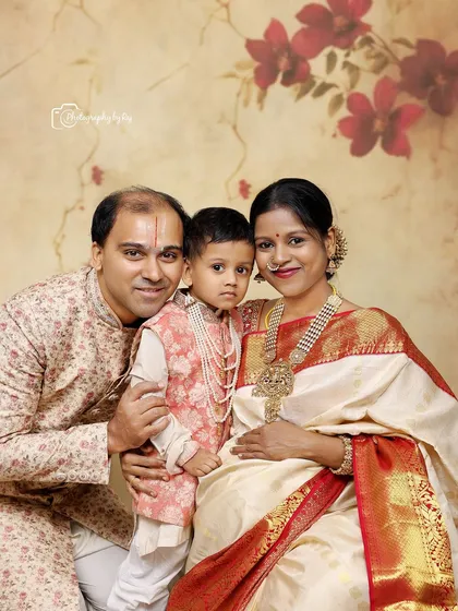 A beautiful traditional family portrait. The mother-to-be is glowing in a white and red saree, surrounded by her husband and son, all in coordinated ethnic wear.