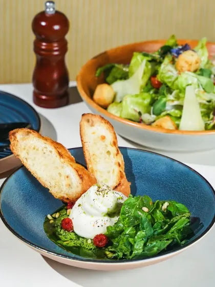A fresh burrata salad with pesto, greens, and toasted bread, served in a beautiful blue ceramic bowl. The bright, hard light creates a clean and contemporary feel, perfect for a modern cafe menu.