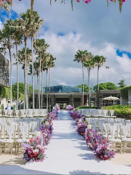 A beautiful outdoor wedding aisle lined with purple and pink flowers, leading to a modern ceremony space framed by palm trees.