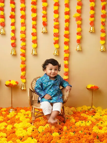 A little boy dressed in a traditional blue kurta for a Ganesh Chaturthi themed shoot. The backdrop of marigold flowers and bells creates a vibrant and festive atmosphere.