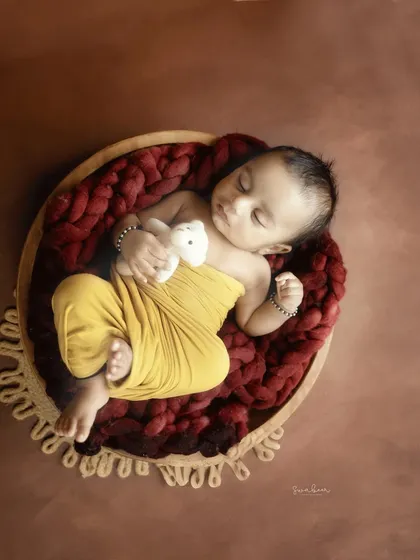 Cuddling a teddy bear, this baby sleeps in a wooden bowl lined with a deep red, chunky knit blanket. The earthy brown background complements the natural tones of the setup.