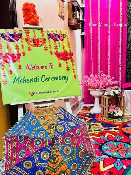 The entrance to a colorful Mehendi/Dholki party, featuring a welcome sign, a traditional umbrella, and a glimpse of the vibrant decor inside.
