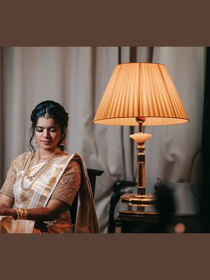 An elegant, moody portrait of a bride in a traditional white and gold saree. The warm lamplight creates a serene and timeless atmosphere.