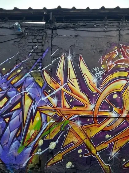 Two distinct graffiti styles side-by-side. On the left, a purple and yellow piece with sharp, aggressive lines, and on the right, an orange and yellow piece with a more rounded, flowing style.