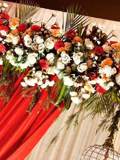 A detailed shot of a floral header with red and peach roses, white carnations, and tropical leaves against a bold red drape. The textures and colors work together to create a rich, celebratory look.