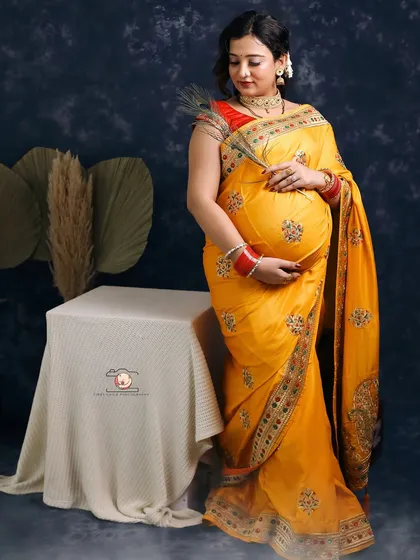 Draped in a vibrant yellow embroidered saree, this mother-to-be radiates joy. The peacock feather she holds is a beautiful symbol of grace and new life, adding a traditional touch to this studio portrait.