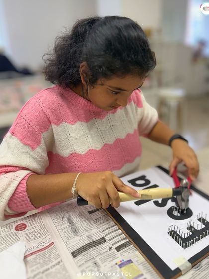 A young artist carefully hammers nails into her nameplate board, focused on getting the placement just right.