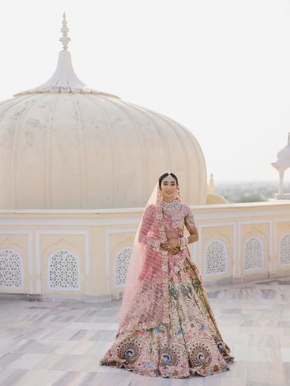 A full-length bridal portrait on a palace rooftop. The bride stands elegantly in her pastel lehenga, with the architectural dome creating a perfect, symmetrical backdrop.