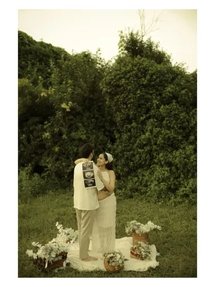 The story begins with this first picture. Here, the couple shares a look of love while holding their baby's sonogram, surrounded by a beautiful picnic setup in a green meadow. It's a perfect pregnancy announcement photo.