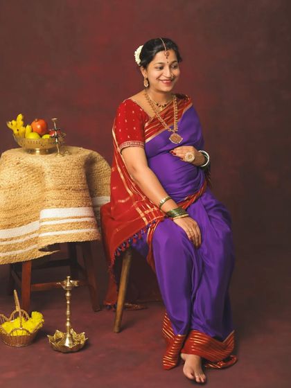 A peaceful and traditional maternity portrait. The mom-to-be is seated gracefully in a classic Nauvari saree, surrounded by props like a brass lamp and fruit basket that evoke a sense of heritage.