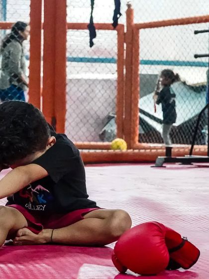 A young boy stretches with his boxing gloves nearby, while other kids train in the background. Our gym is a safe and motivating space for children to learn and grow.