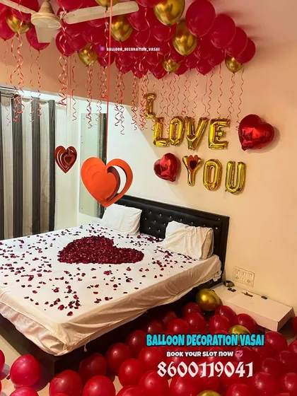 A full view of a bedroom filled with red and gold balloons on the floor and ceiling. The bed is the centerpiece, with a large rose petal heart and "I LOVE YOU" message.