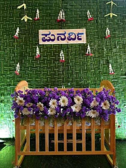 A traditional naming ceremony setup featuring a backdrop of woven coconut leaves (thennai olai). The baby's name is displayed in a custom Kannada script, and the wooden cradle is adorned with purple and white flowers.