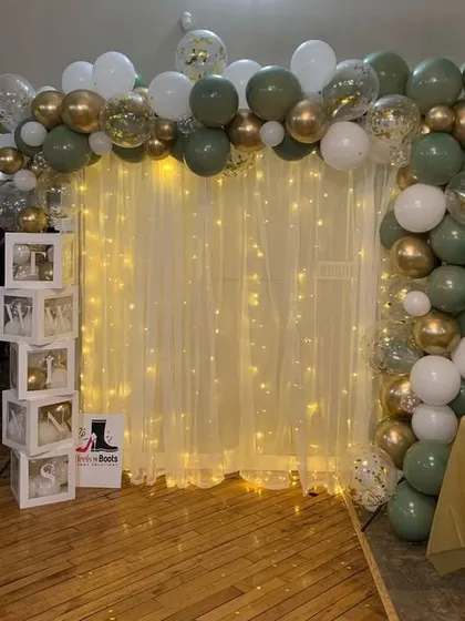 An elegant and simple backdrop solution for any event. This design features a curtain of warm fairy lights behind a sheer white drape, framed by an organic balloon garland in olive green, white, and gold.