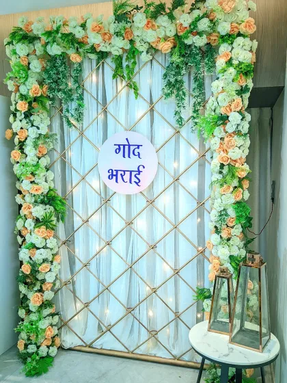 An elegant and simple backdrop for an at-home 'Godh Bharai' ceremony. The design features a lattice frame decorated with peach and white flowers, with fairy lights woven in for a gentle glow.