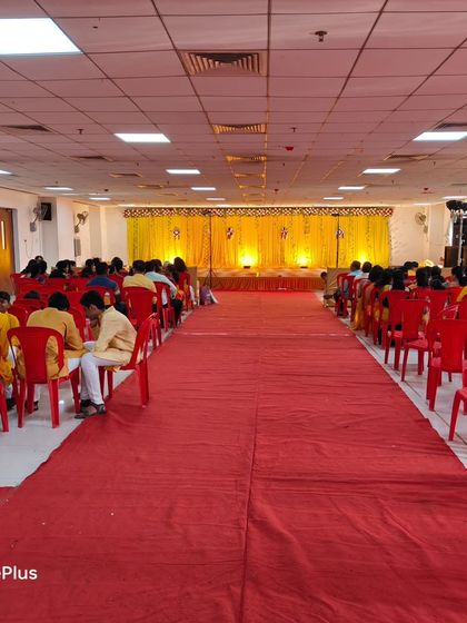 The seating arrangement for a Haldi ceremony in a hall. The red carpet and chairs are set up facing the bright yellow stage.